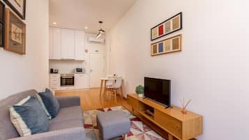 Uma sala de estar moderna com um sofá cinzento, uma televisão e um móvel para TV de madeira. Ao fundo, cozinha com armários brancos e um espaço de jantar com mesa e cadeiras.