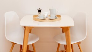 Un coin repas minimaliste avec une table blanche, deux chaises blanches, un plateau contenant une théière, des tasses et un récipient.