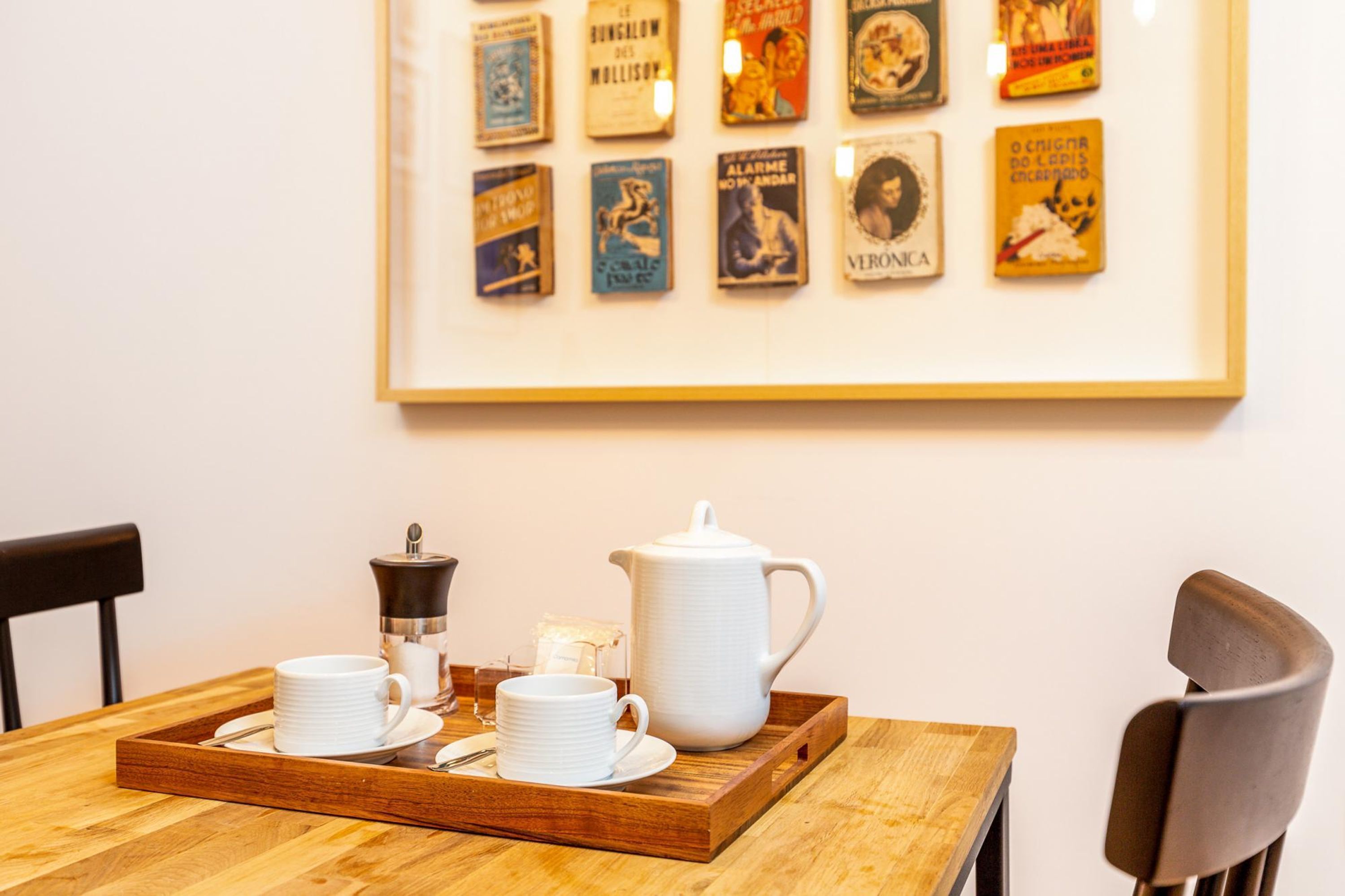 Uma mesa de jantar com uma bandeja contendo um coador de café, xícaras e pires. Uma coleção de livros emoldurada está exposta na parede ao fundo.