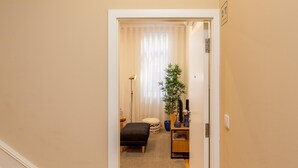 En smal hall leder in till ett rum med ett stort fönster, en krukväxt, en svart pall och ett litet bord i trä.