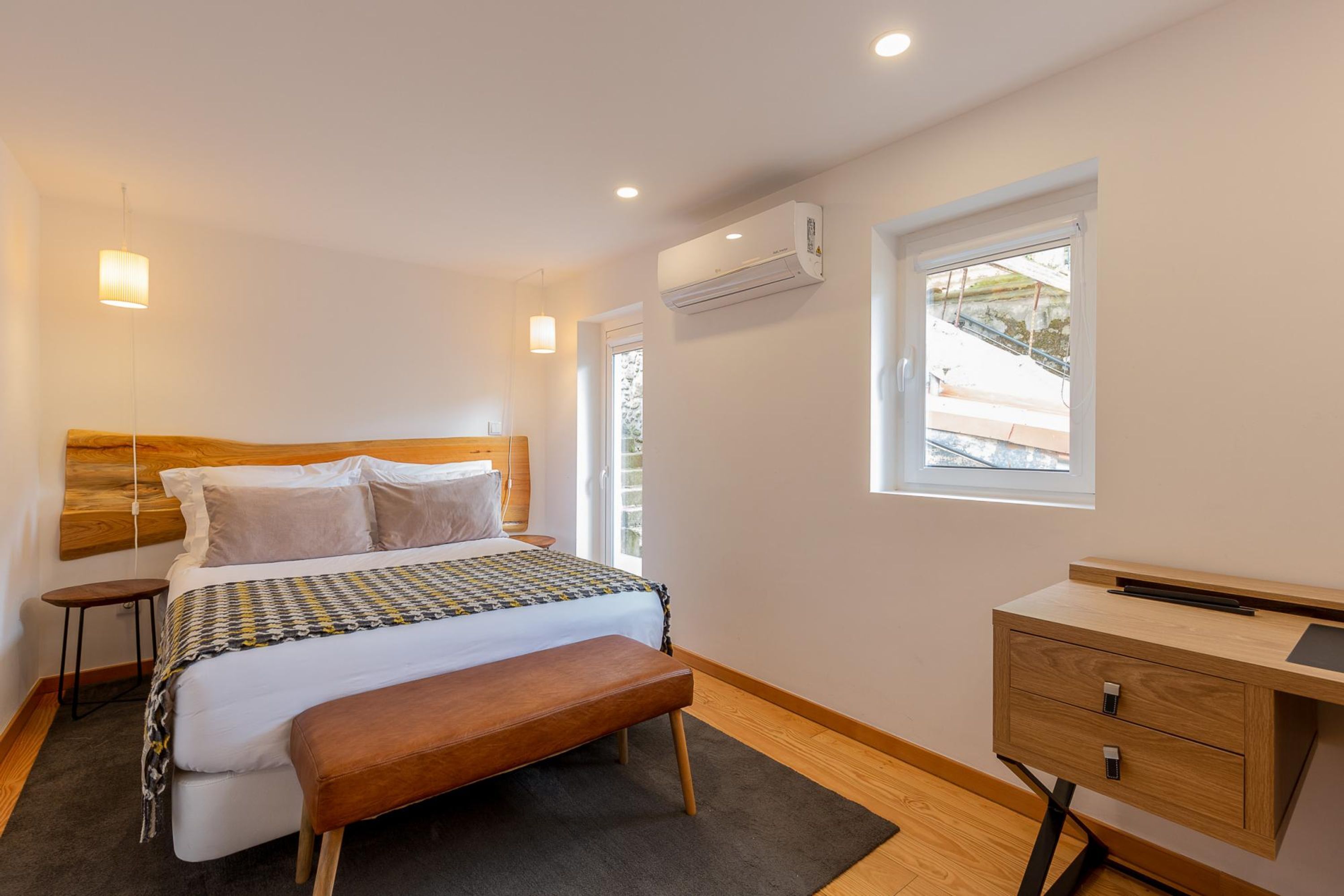 Quarto moderno com cabeceira de madeira, cama com manta estampada, um banco, mesa de cabeceira, escrivaninha com gavetas e um aparelho de ar condicionado.