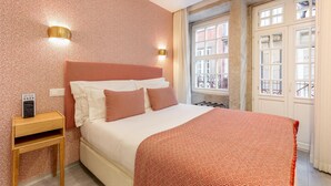 A hotel room with a patterned wall, a bed with white linens and patterned pillows, a bedside table with a lamp, and a window with a view of buildings. - Ribeira Porto 1 in Oporto (Porto)
