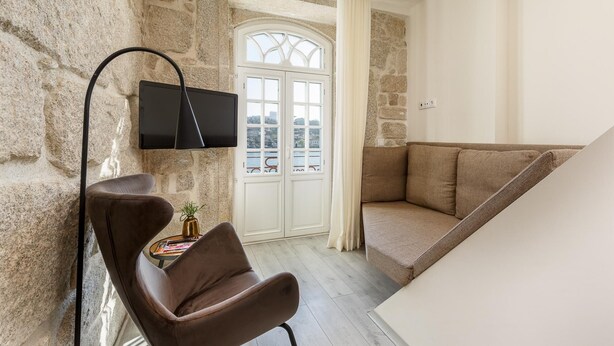 A modern living room with a stone wall, a flat-screen TV mounted on the wall, a brown leather chair, a small table with a plant, and a beige sofa. - Ribeira Porto 1 in Oporto (Porto)