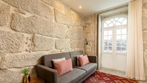 Une pièce avec un mur en pierre, un canapé gris agrémenté de coussins rouges, une petite table ronde avec une plante et un tapis coloré.