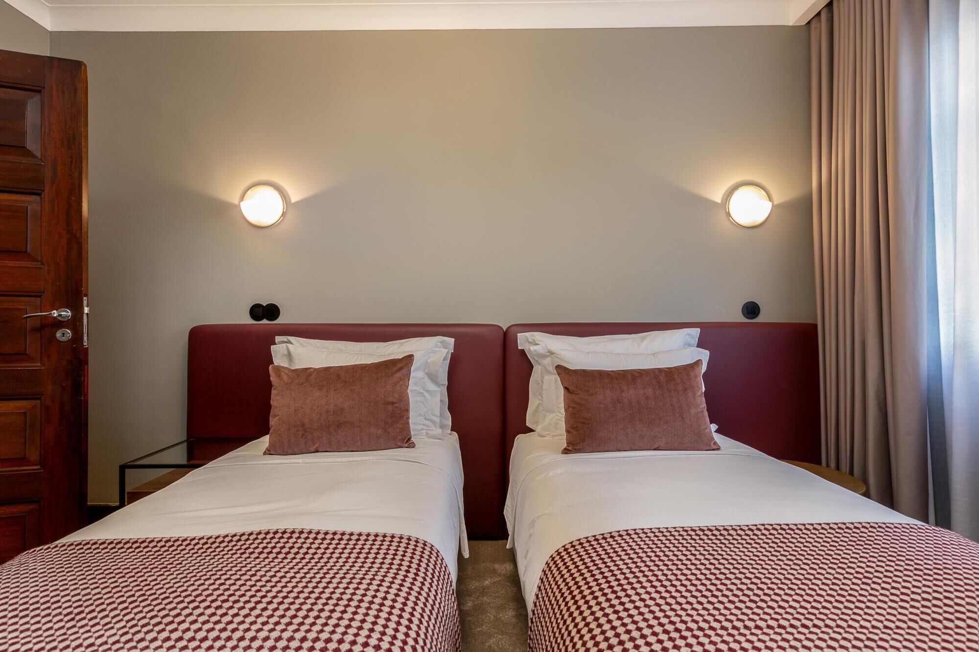 Hotellihuoneessa on kaksi sänkyä, joissa on valkoiset lakanat ja ruskeat tyynyt sekä ruudullinen punavalkoinen peitto. Kummankin sängyn yläpuolella on seinään kiinnitetyt valaisimet, ja vasemmalla puolella on puinen ovi.
