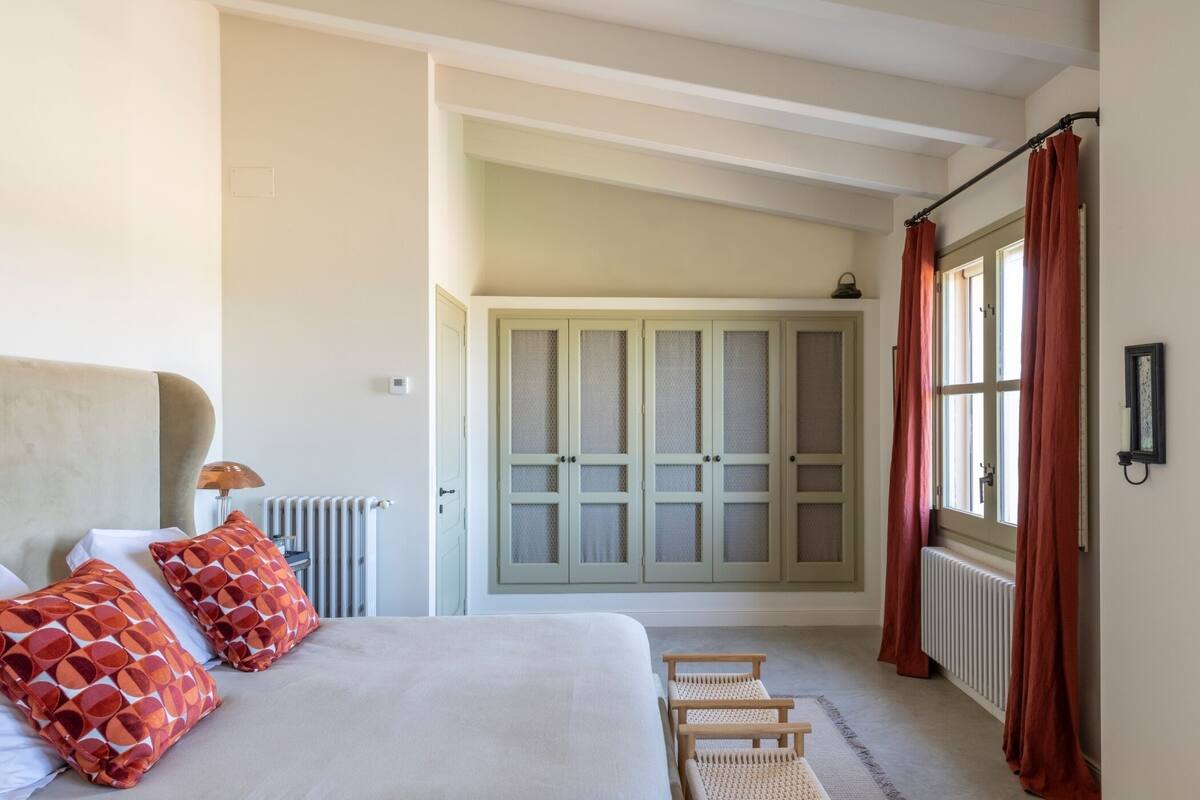 A bedroom with a bed, pillows, a radiator, a window with curtains, and a wardrobe.