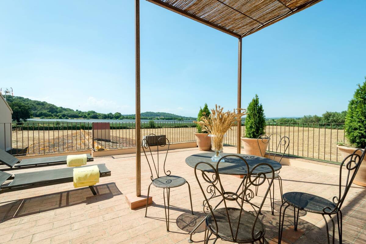 A terrace with a table and chairs, a sun lounger, and a view of a lake and hills.