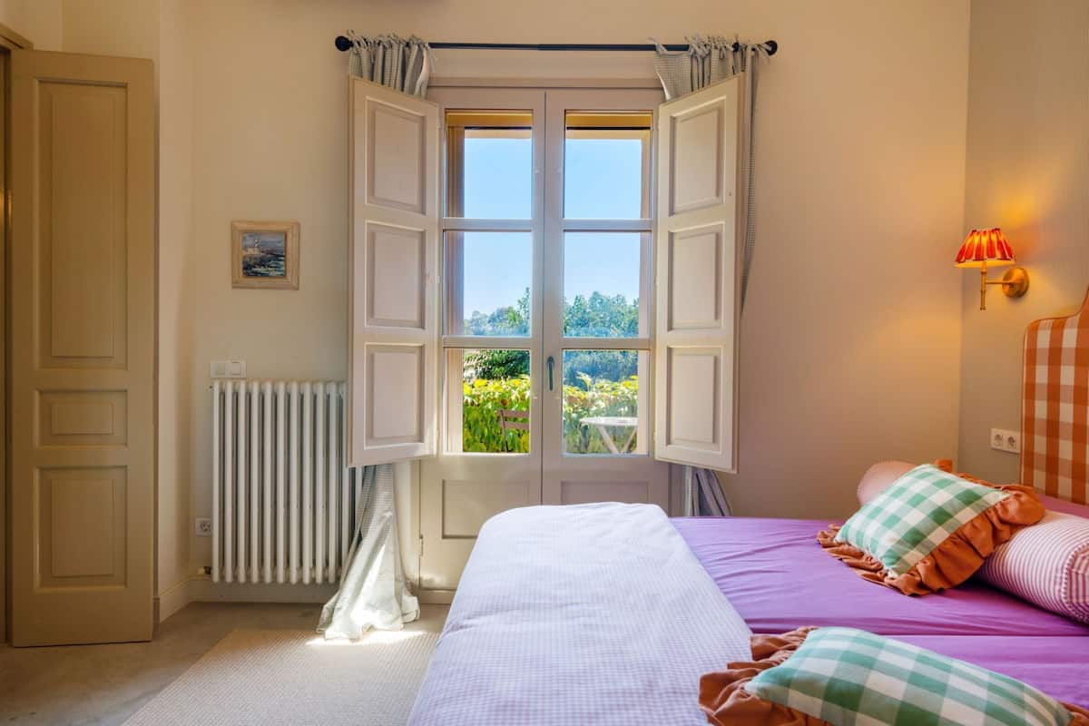 A bedroom with a large window, a radiator, a bed with checkered pillows, and a wall-mounted lamp.