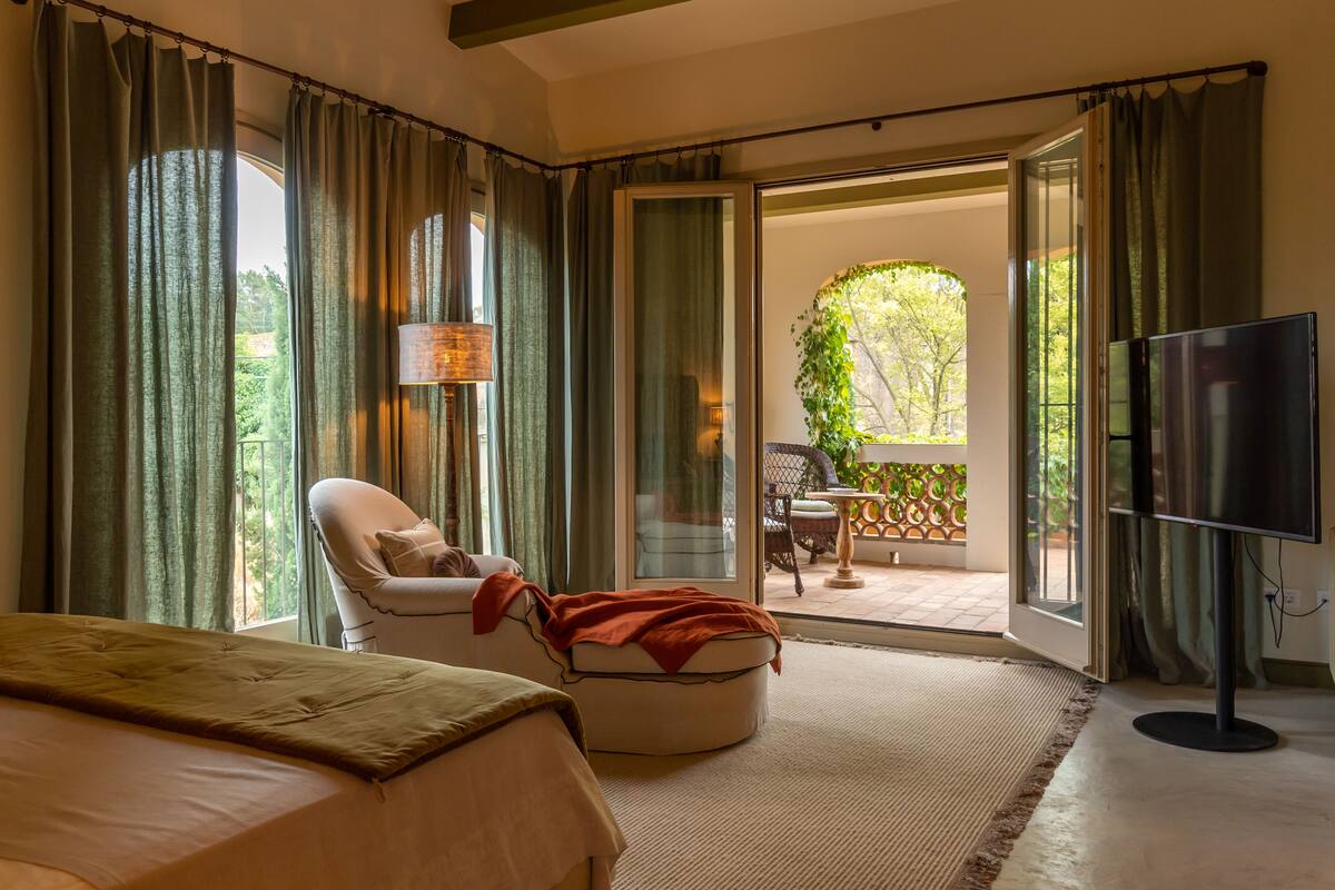 A bedroom with a bed, a chair, a TV, and a sliding glass door leading to an outdoor area with a table and chairs.