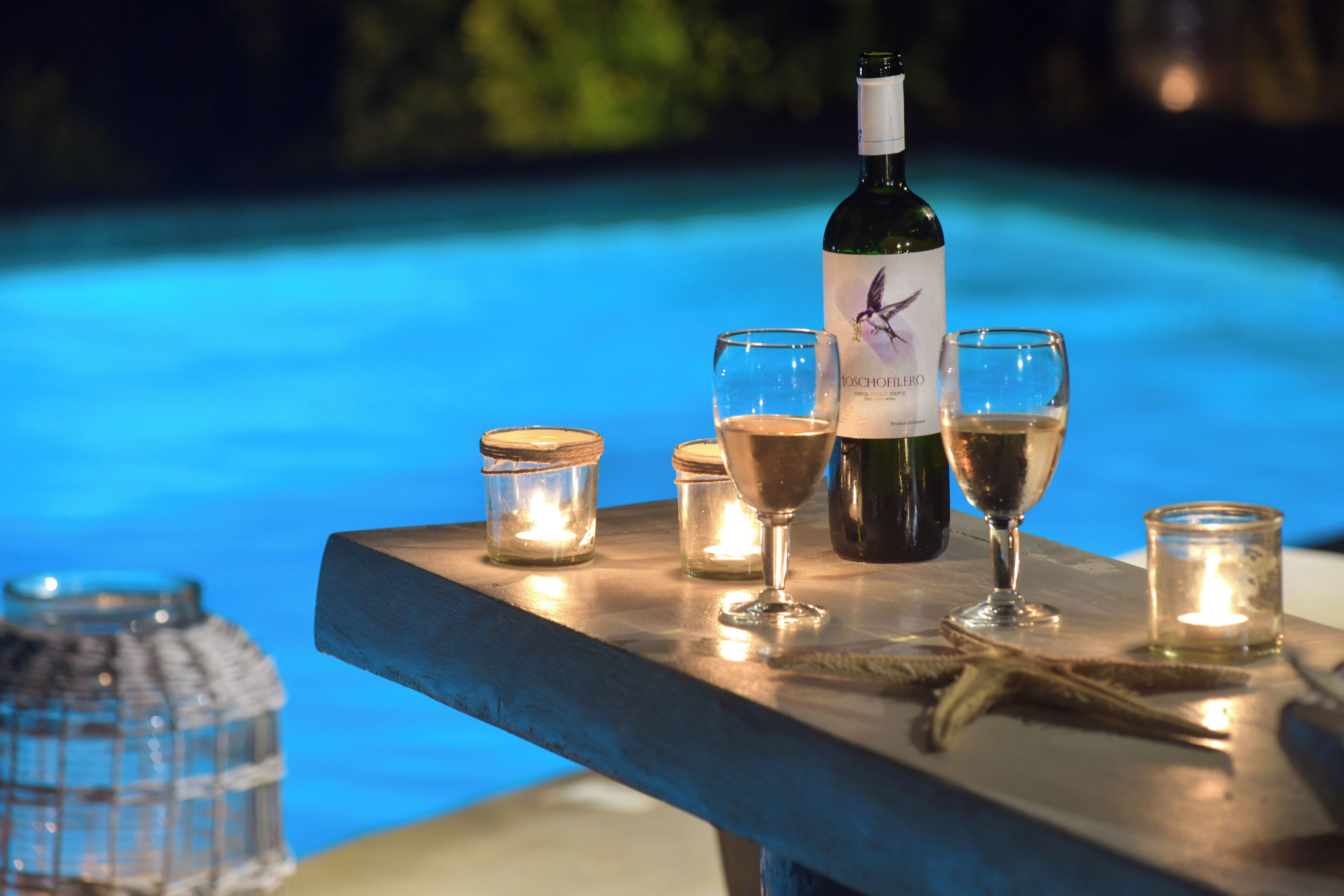 A bottle of wine, two glasses, and candles on a table by a pool.