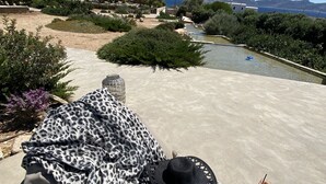 Una tumbona con una manta de estampado de leopardo, un sombrero negro y chanclas sobre una superficie de hormigón con una piscina y vegetación al fondo.