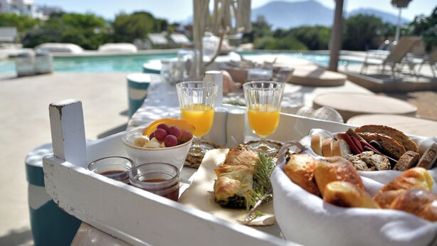 Bandeja de desayuno con pasteles, frutas y vasos de jugo junto a una piscina con vista a la montaña.