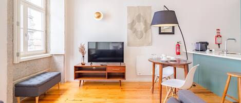 A modern living room with a flat-screen TV, a bench, a sofa, a dining table with chairs, a coffee machine, and a fire extinguisher.