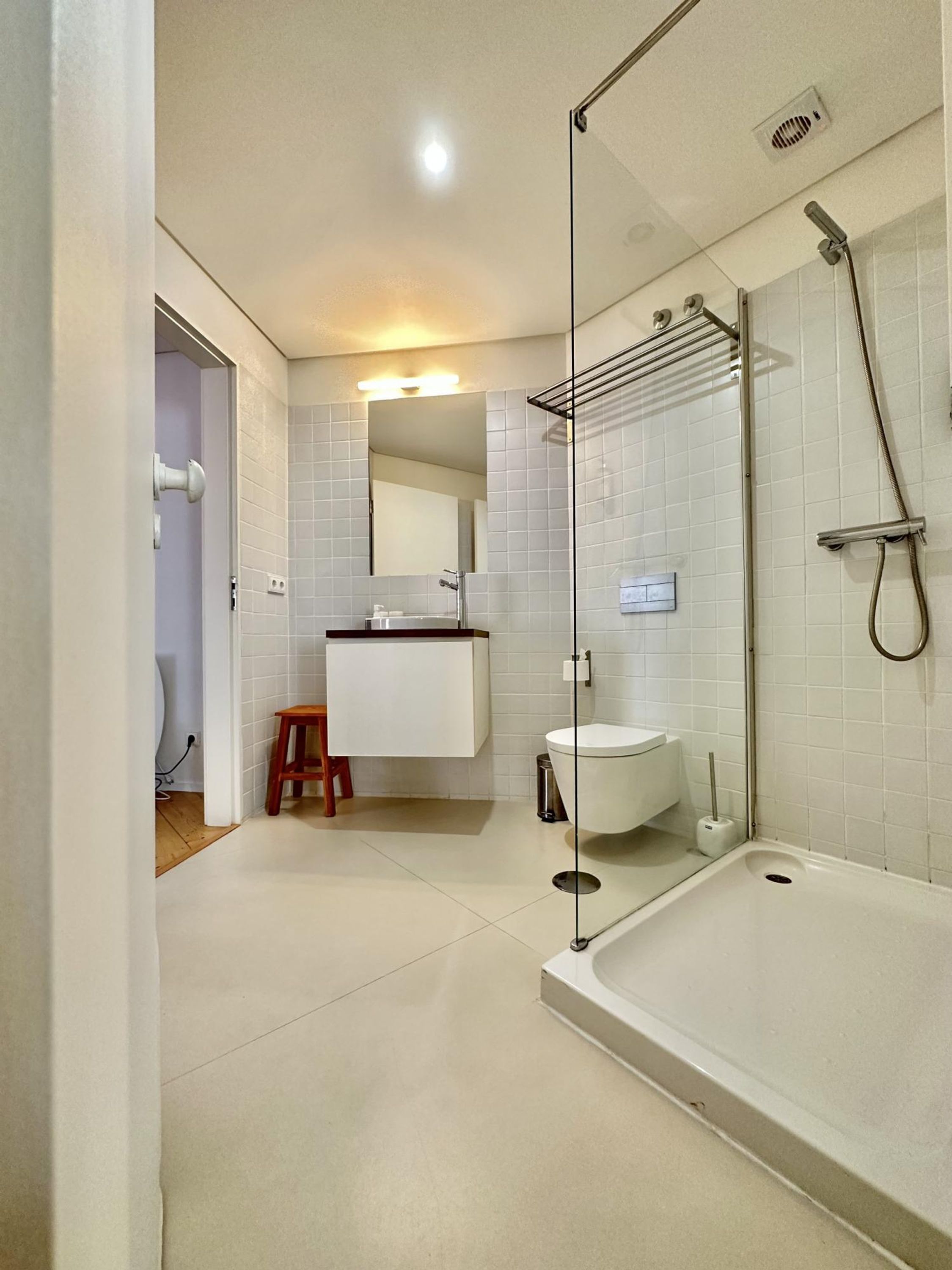 A modern bathroom with a glass shower enclosure, a white sink, and a wooden stool.