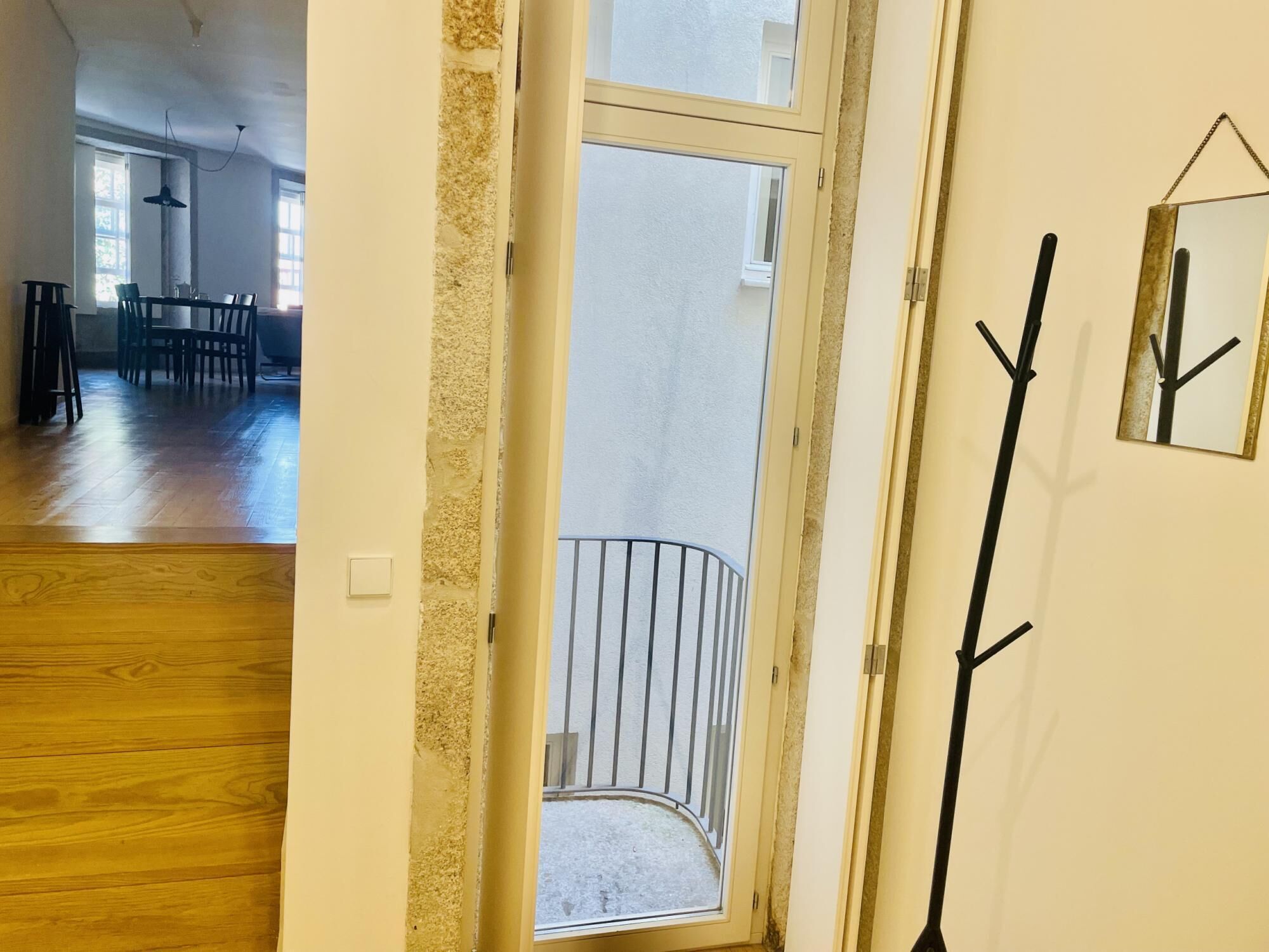 Eine geöffnete Tür, die auf einen Balkon mit Geländer führt, ein Essbereich mit Stühlen und einem Tisch sowie ein Garderobenschrank mit Spiegel.