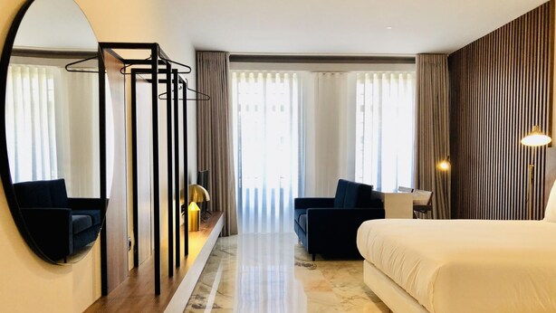 A modern hotel room with a large bed, a seating area with a sofa and armchairs, a wooden console table, a large mirror, and a view of the outdoors through large windows. - Downtown 2A in Oporto (Porto)