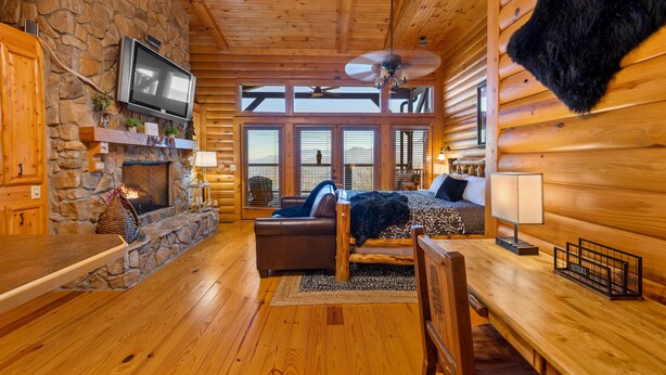 Ein Holzblockhaus-Interieur mit Kamin aus Stein, Holzwänden, einem an der Wand montierten Fernseher, einem Deckenventilator und einem Bett mit Blick auf die Berge durch große Fenster.