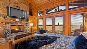 Ein Holzblockhaus-Interieur mit Kamin aus Stein, einem Bett mit dunkler Decke und einem an der Wand montierten Fernseher.