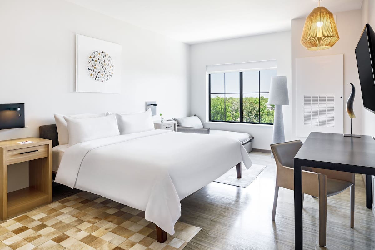 A modern hotel room with a large bed, a desk, a chair, a TV, and a window with a view of greenery.