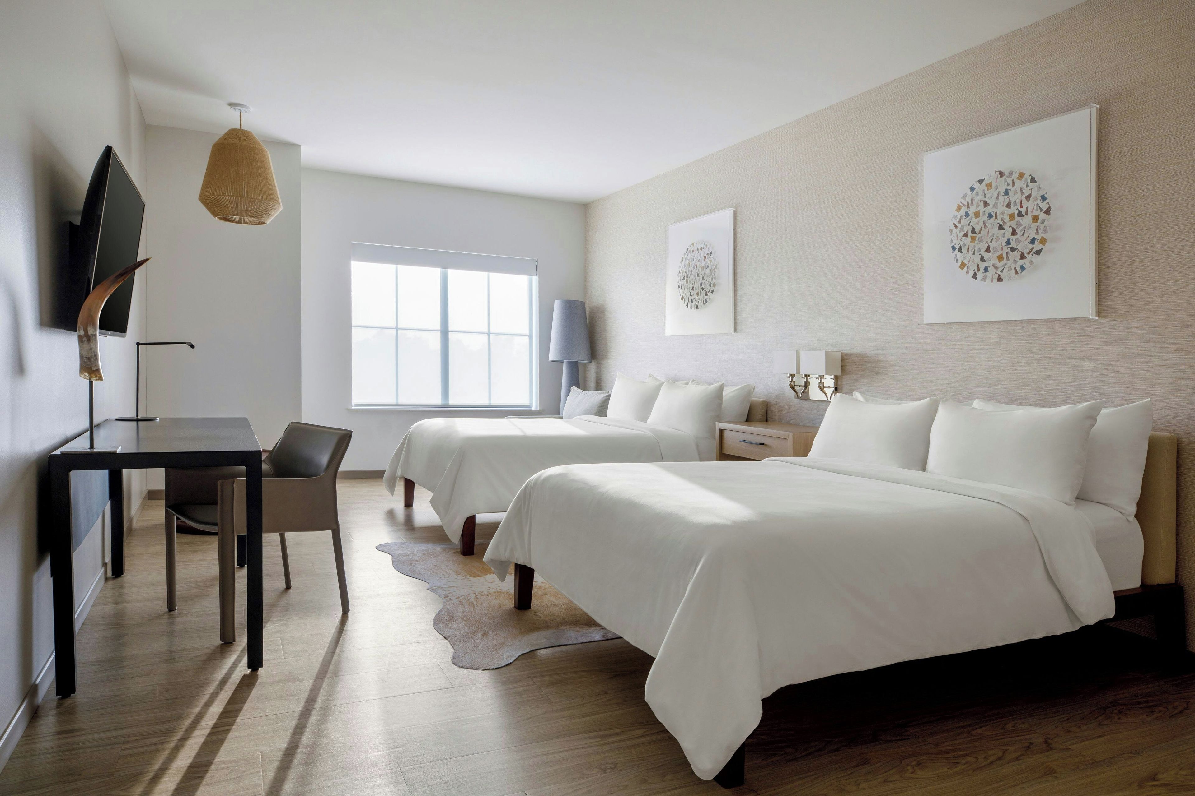 A modern hotel room with a desk, two beds, a large window, and artwork on the walls.