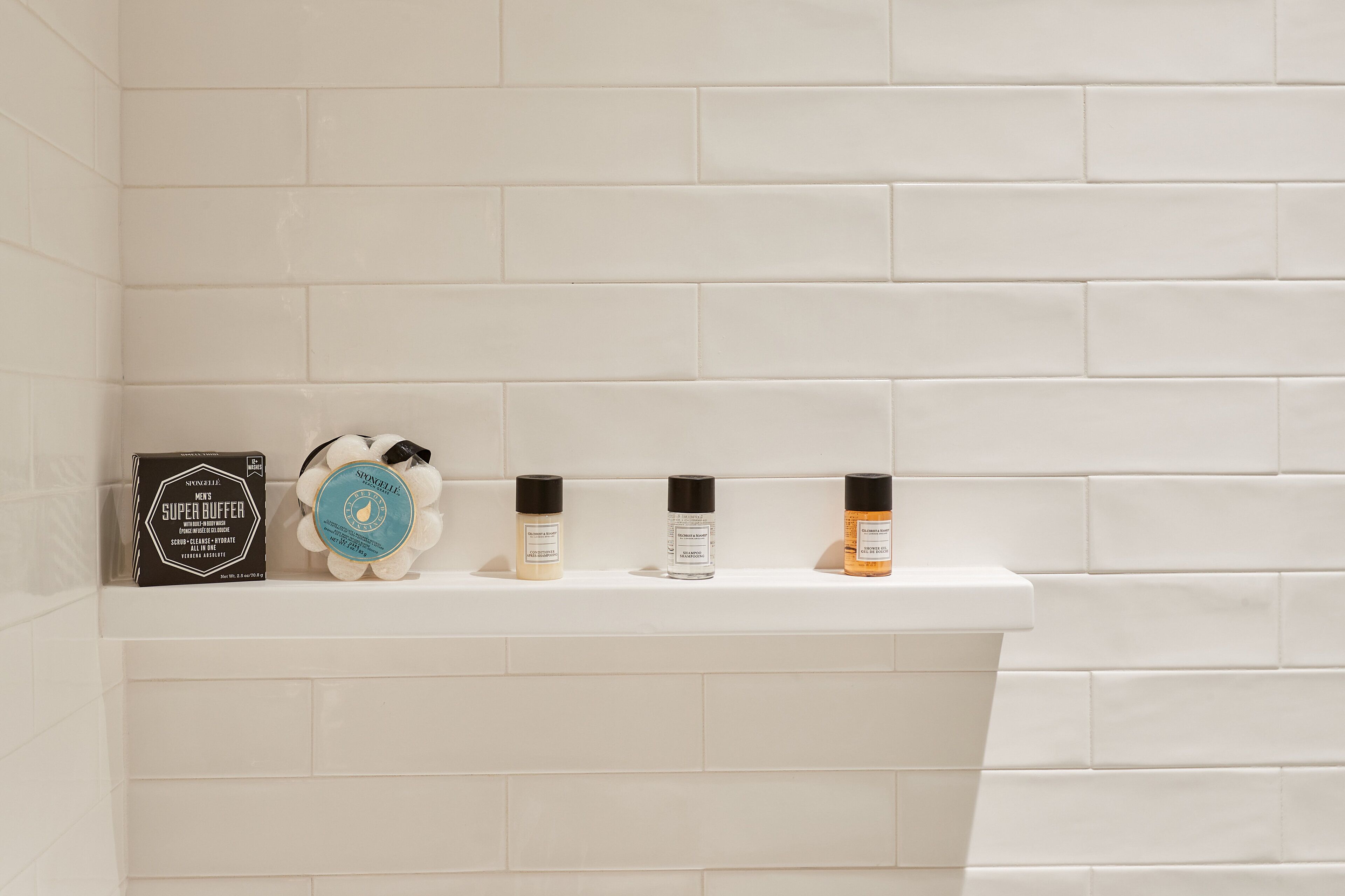 A bathroom with white tiles and a shelf holding three bottles of toiletries.