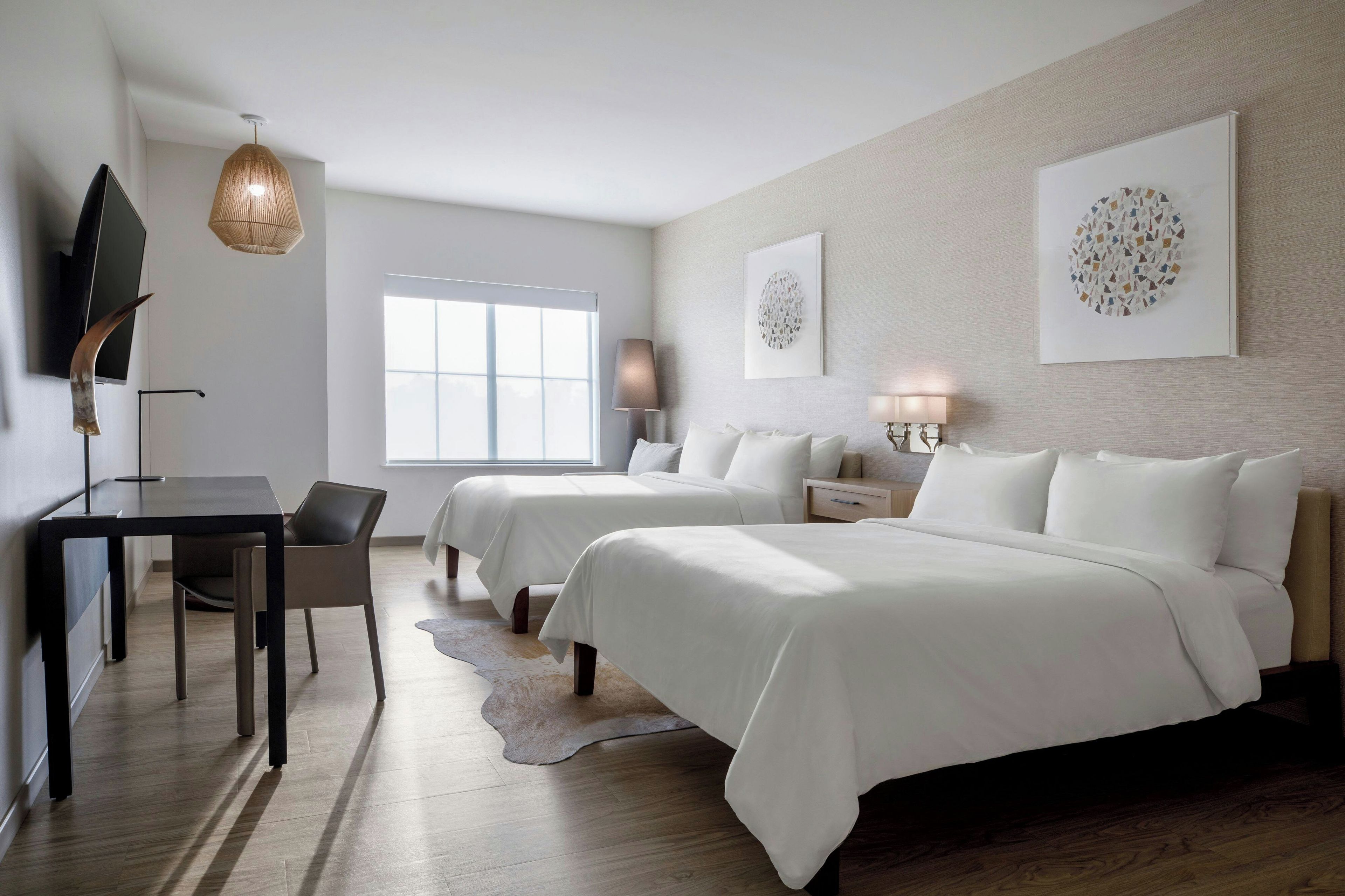 A modern hotel room with a desk, two beds, a large window, and artwork on the walls.