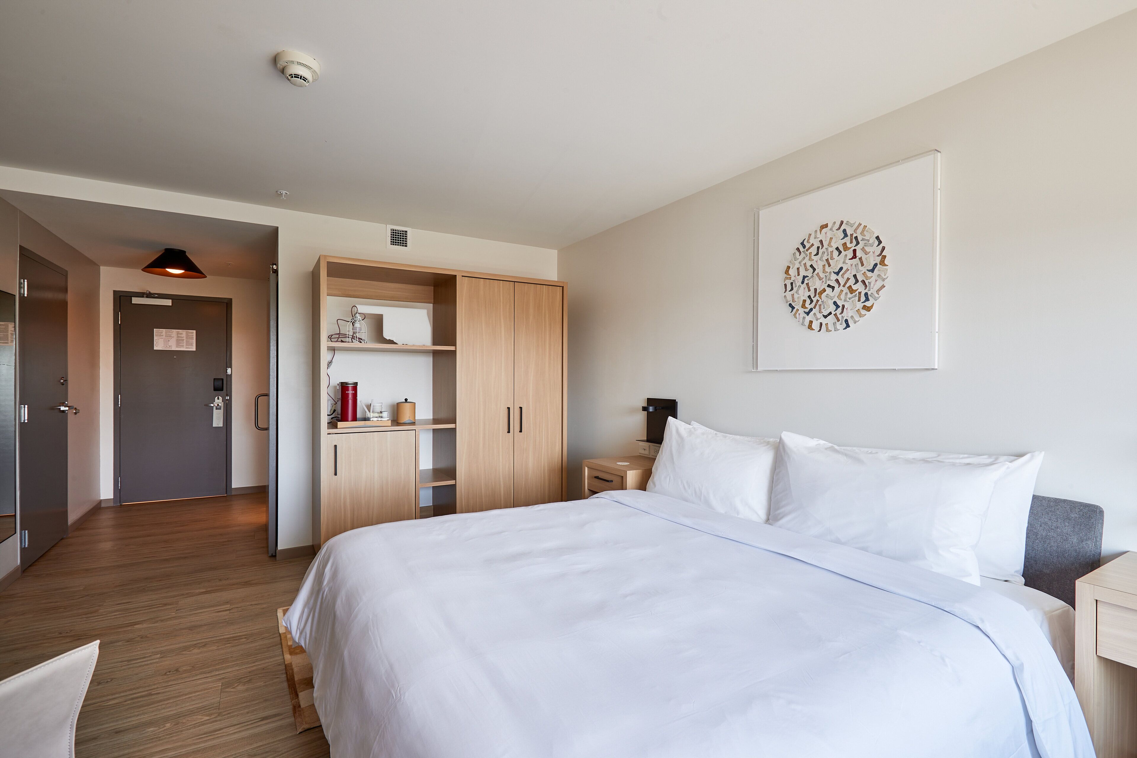A hotel room with a large bed, wooden wardrobe, a small desk, and a wall-mounted artwork.