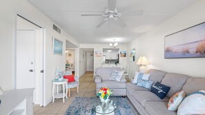 Uma sala de estar com sofá, mesa de centro com flores, ventilador de teto e cozinha ao fundo.