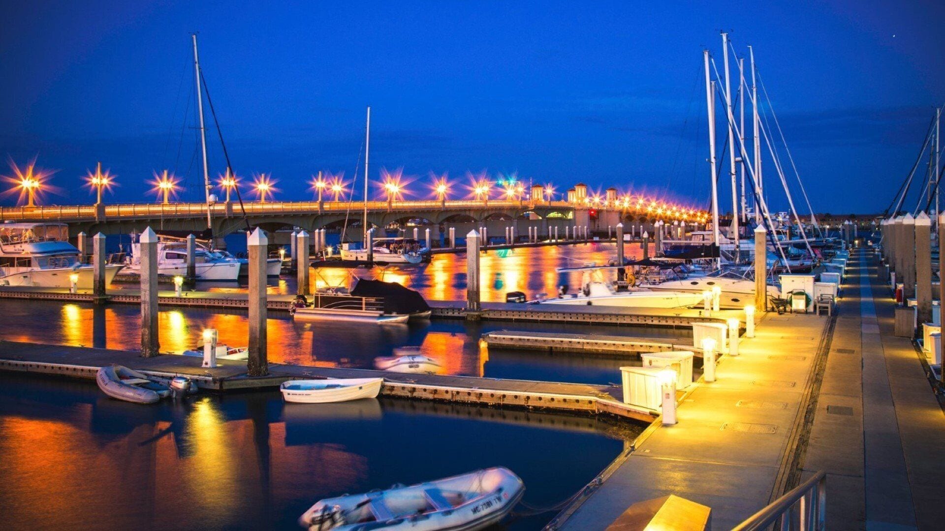 A marina with boats docked, a bridge overhead, and a well-lit waterfront.
