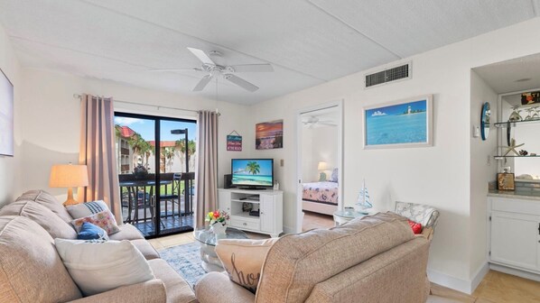 Een woonkamer met een bank, televisie en een balkon met uitzicht op palmbomen en een gebouw.