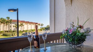 Un balcon avec une table dressée pour deux personnes, agrémentée d’une bouteille de vin et de deux verres à vin. Sur la table, on trouve également un pot de fleurs.