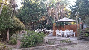 Un espace extérieur aménagé avec une table et des chaises sous une tonnelle, entouré d’une végétation luxuriante et d’une clôture en bois.