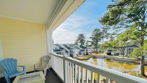A covered porch with Adirondack chairs, a small table, and a view of a lake with houses in the background.
