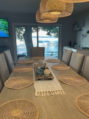 Uma área de refeições com uma mesa comprida, jogos americanos trançados e vista para um cais e para a água.