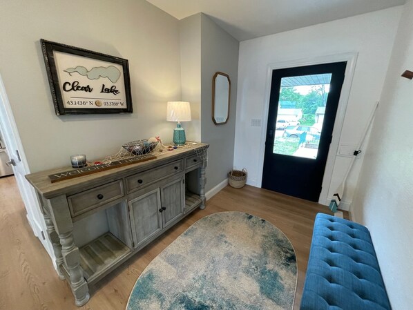 Quarto com cômoda de madeira, um quadro emoldurado, luminária, um banco azul e vista por uma porta.