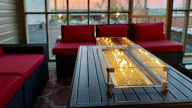 Un rincón al aire libre acogedor con una mesa larga y cojines rojos, con vistas a una puesta de sol a través de amplias ventanas.