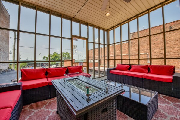 Una habitación luminosa con ventanas amplias, mobiliario de mimbre con cojines rojos y una mesa con cubierta de vidrio.