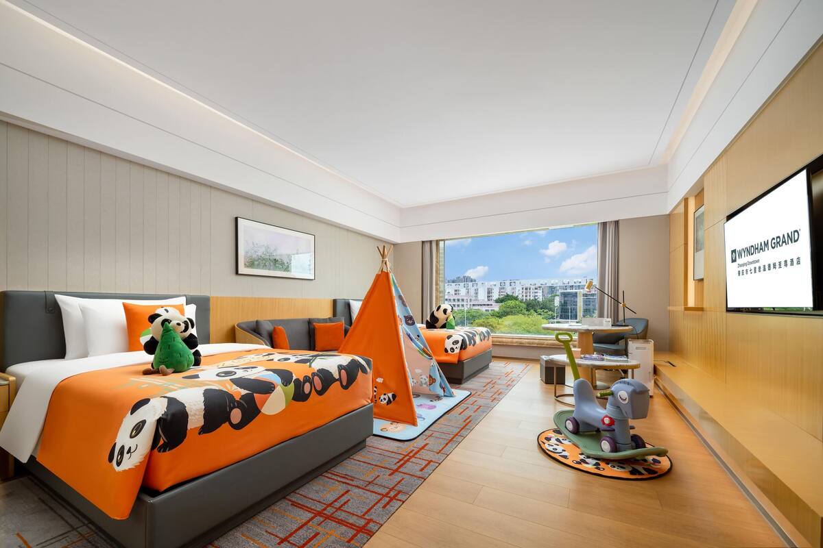 Una habitación de hotel moderna con una cama grande, un pequeño sofá, una zona de juegos con una tienda tipi y juguetes, y un ventanal con vistas a la ciudad.