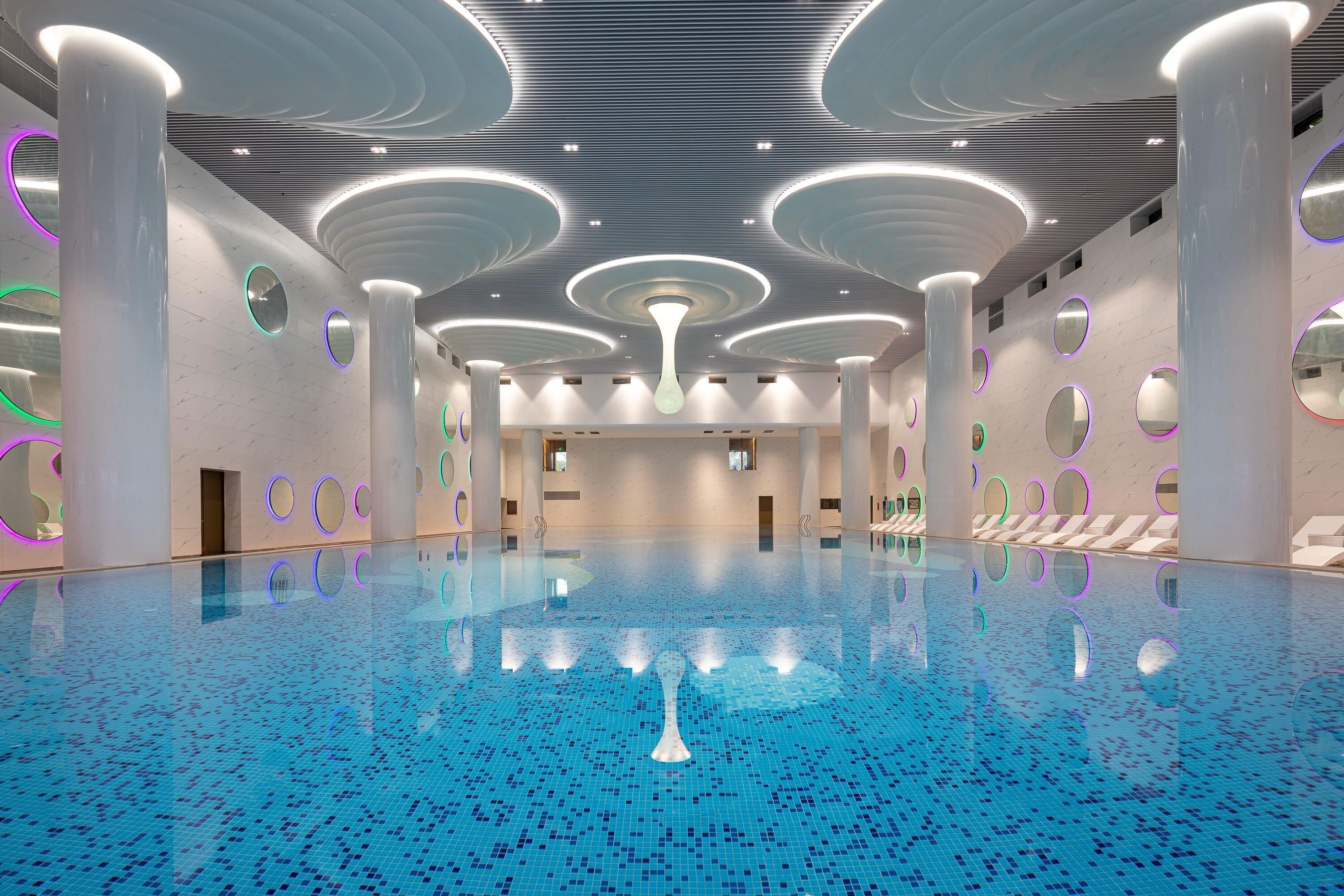 Una amplia piscina interior con un diseño moderno, con luces circulares en el techo y una lámpara central.