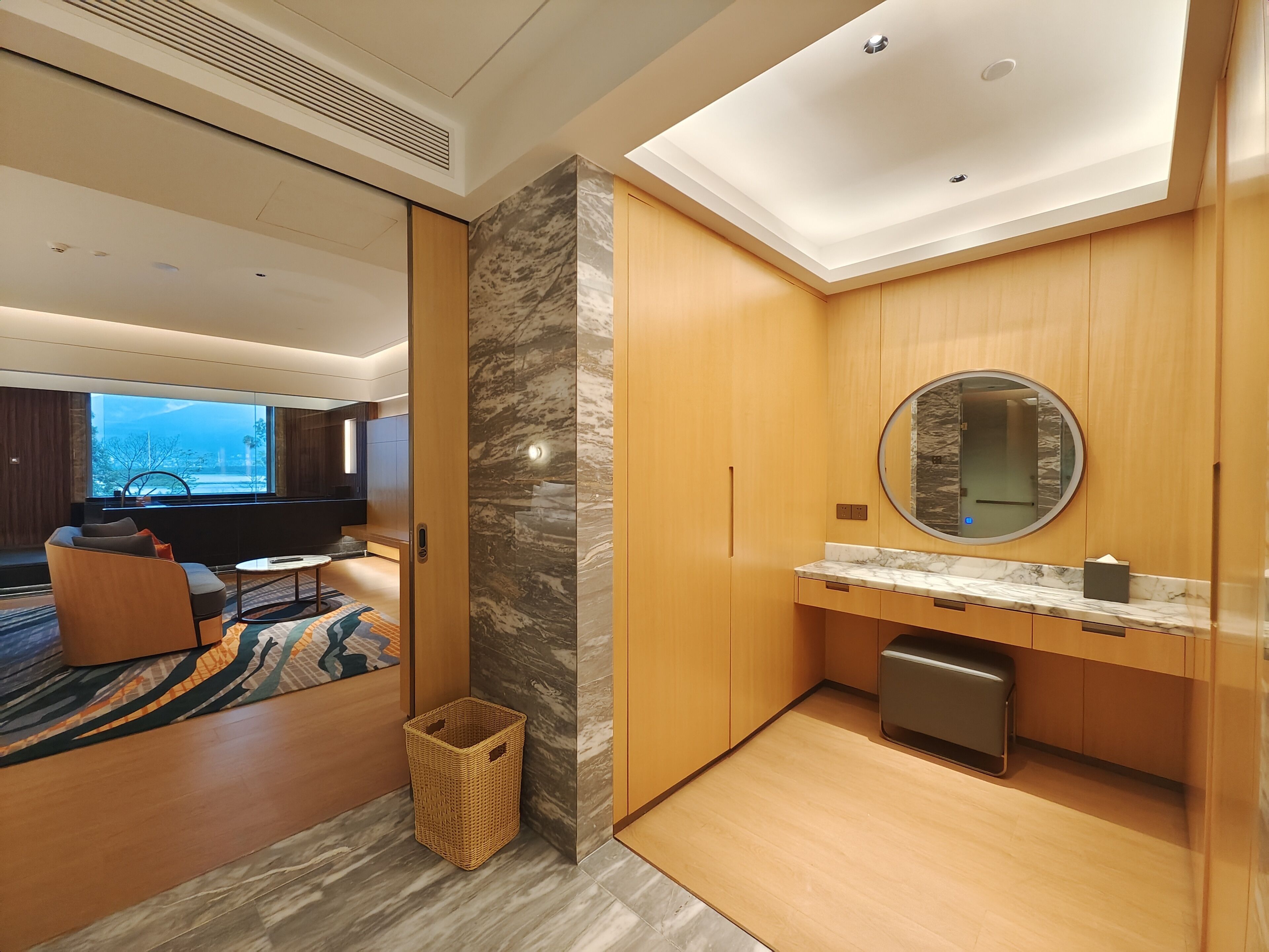 Une chambre d’hôtel moderne dotée d’une grande fenêtre, d’un coin salon confortable, d’un mur décoratif en marbre et d’une coiffeuse en bois avec un miroir.