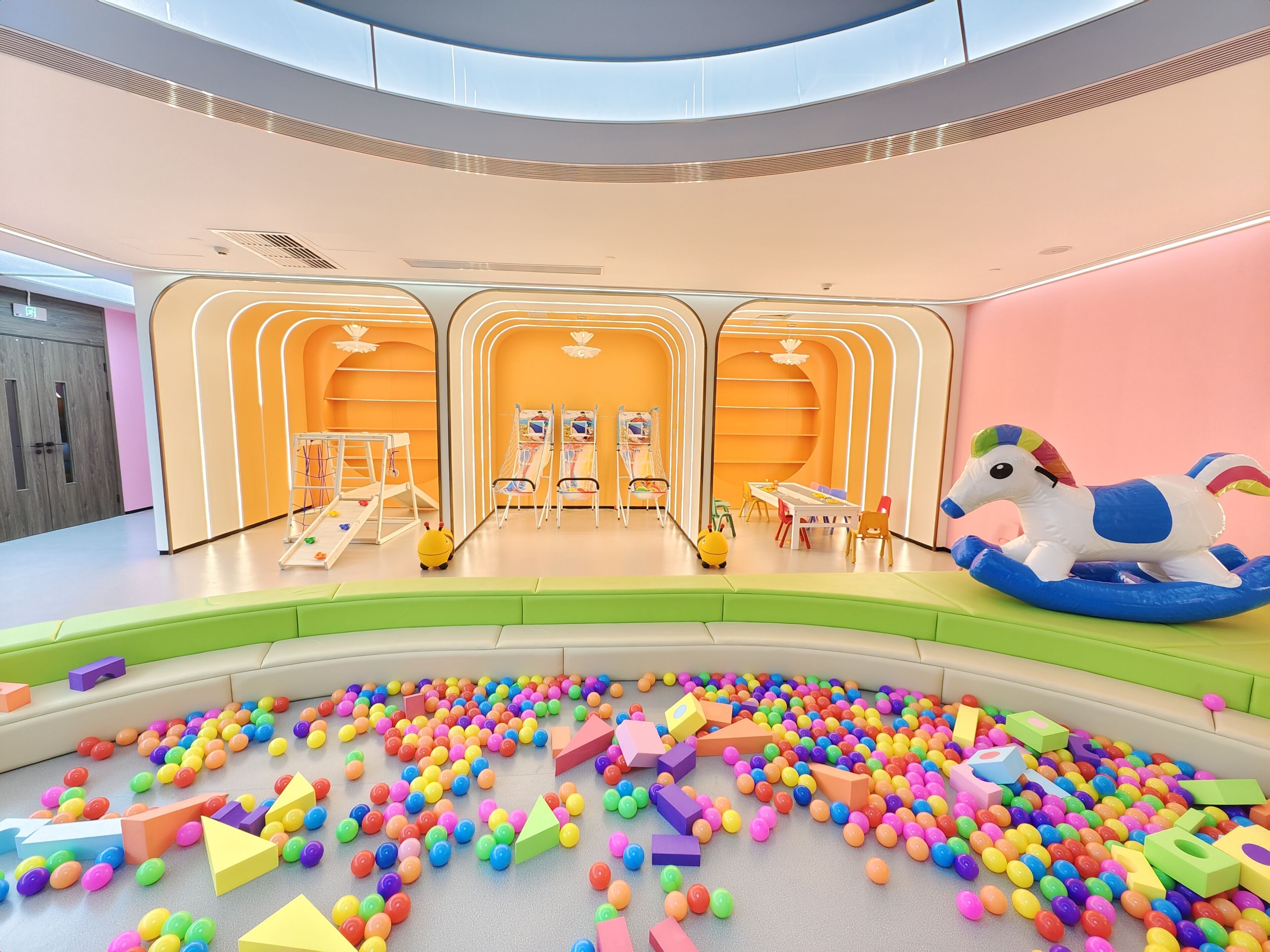 A brightly lit indoor play area with colorful balls and foam shapes on the floor, a carousel, and a playpen with a toy horse.