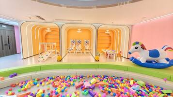 A brightly lit indoor play area with colorful balls and foam shapes on the floor, a carousel, and a playpen with a toy horse.