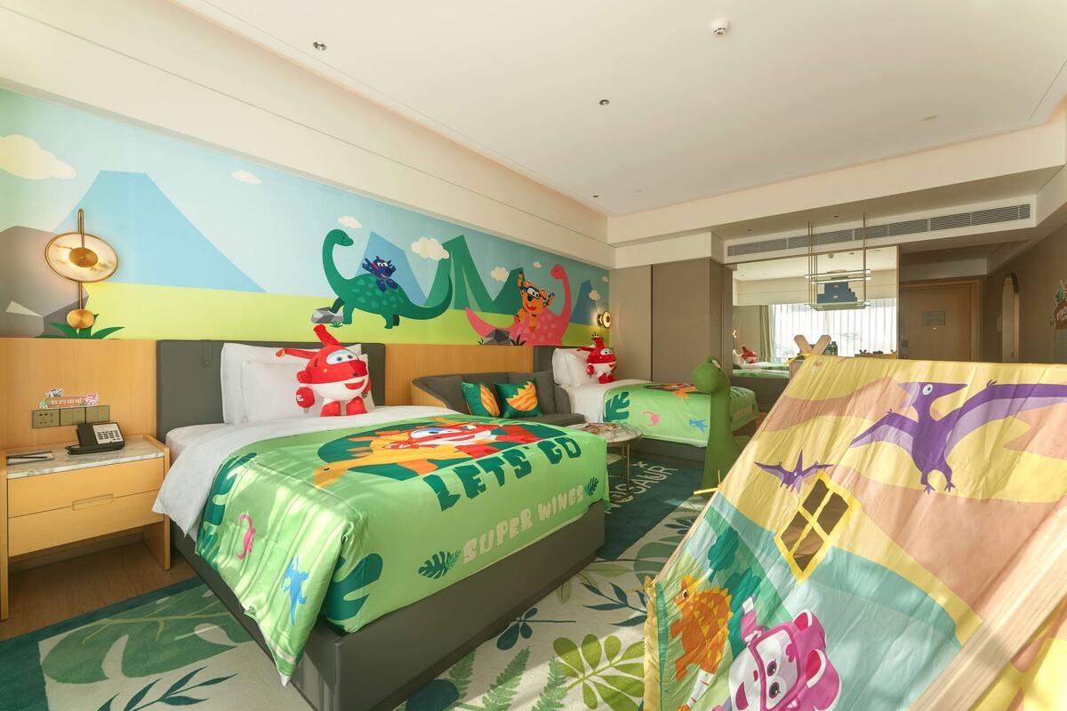 Una habitación infantil con un mural, una cama con colcha de 'Let's Go Super Wings', un escritorio con lámpara y una ventana con persianas.