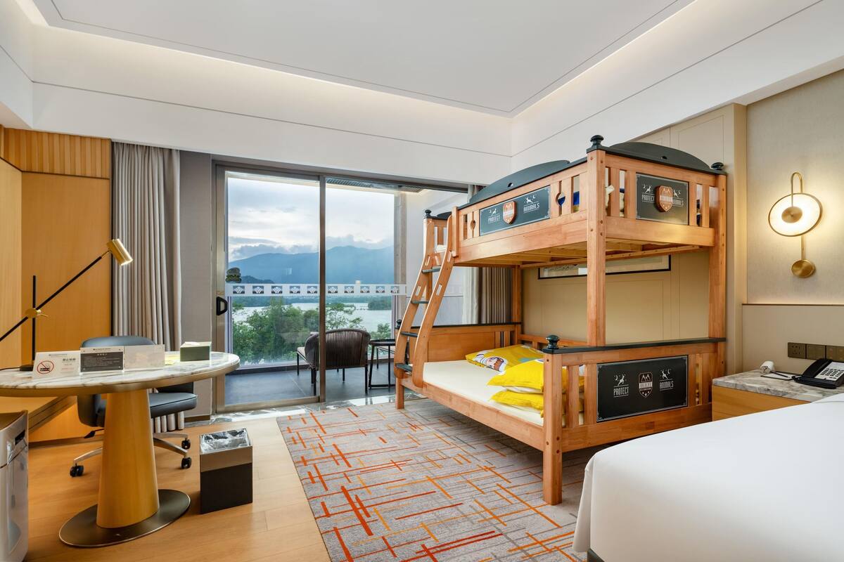 Una habitación de hotel con literas, un escritorio con silla, una mesita, un cesto de basura y vistas a las montañas a través de amplias ventanas.