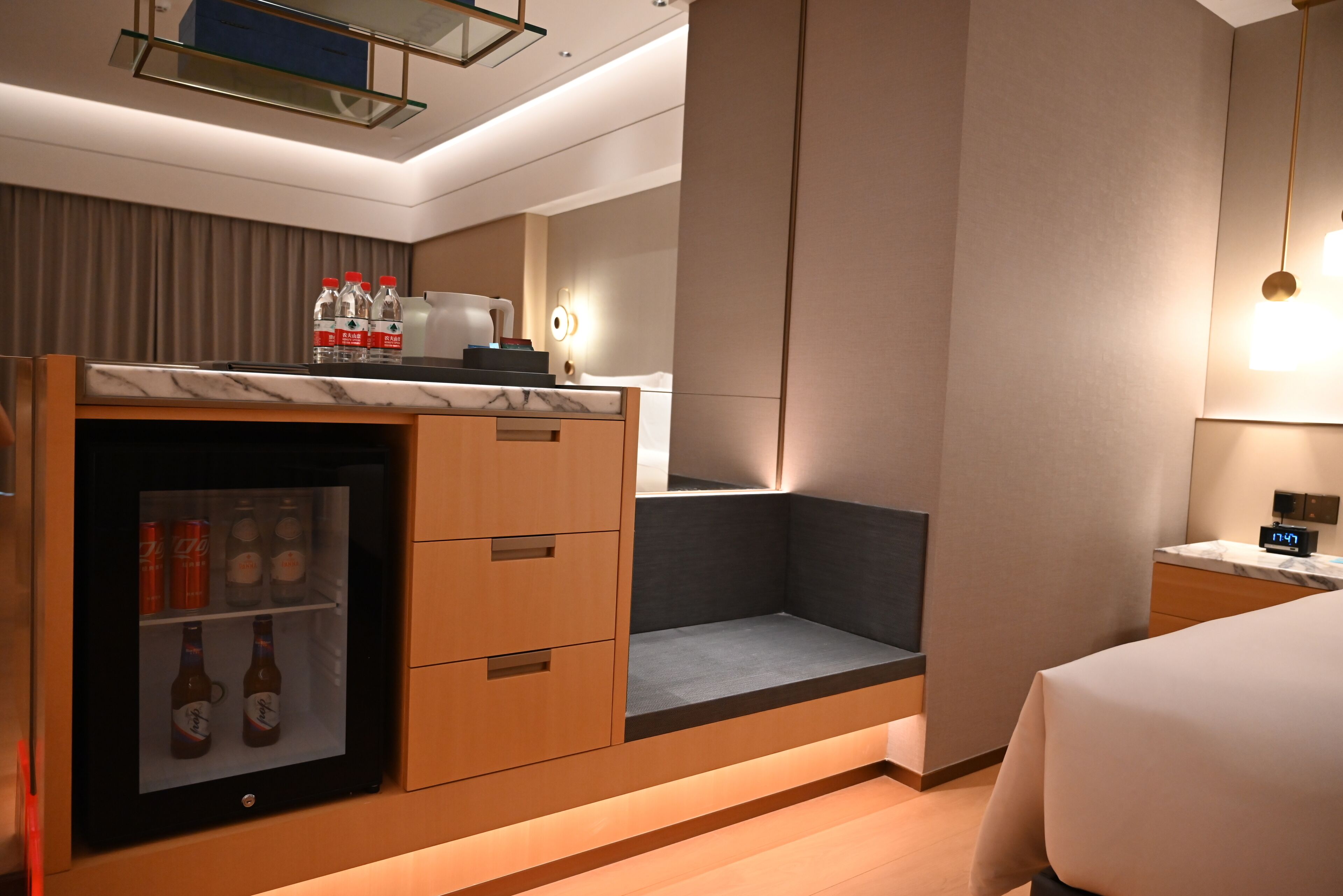 A compact hotel room with a minibar, a bed, a desk, and a seating area.