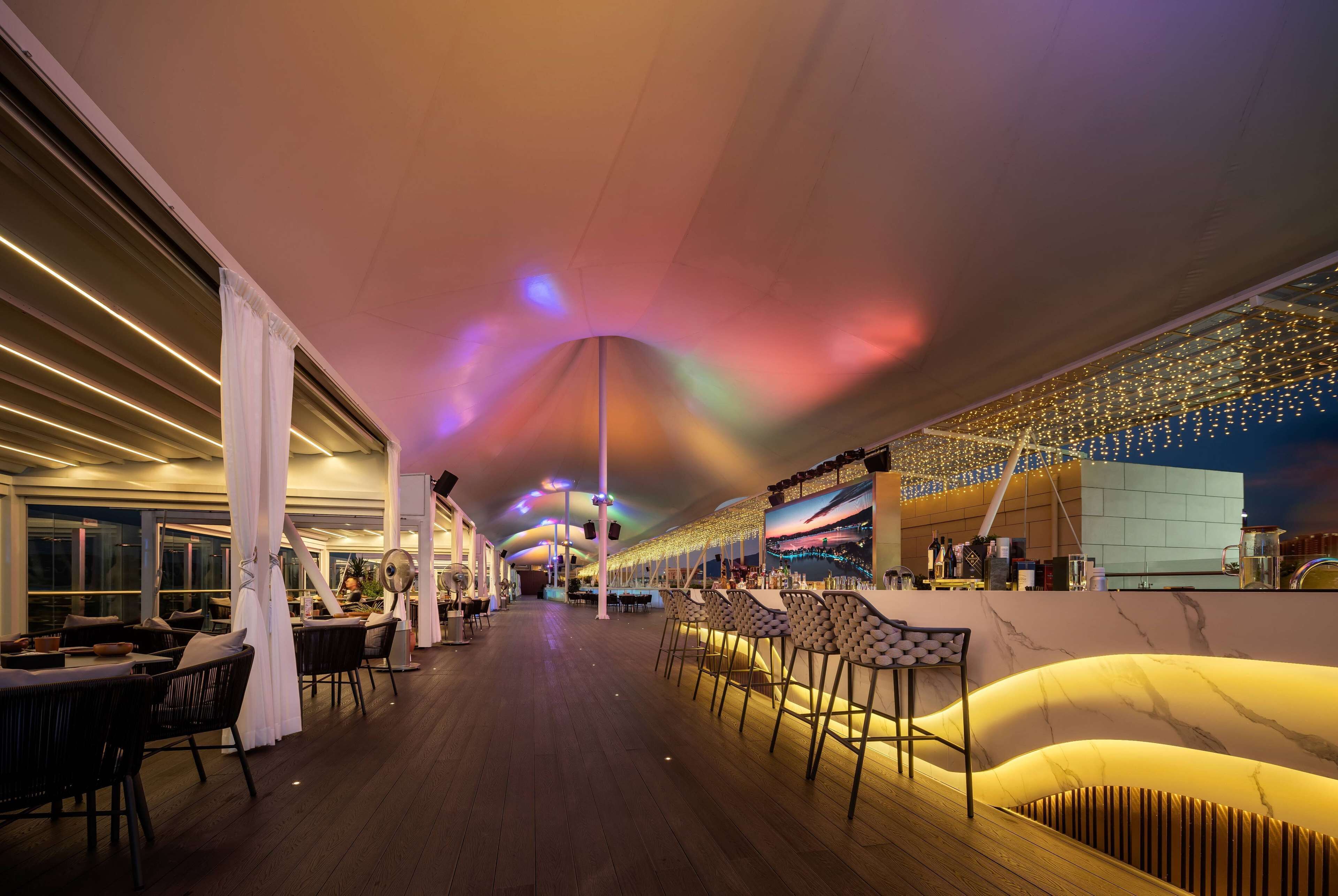 A spacious indoor area with a long bar, seating arrangements, and a view of the sea.