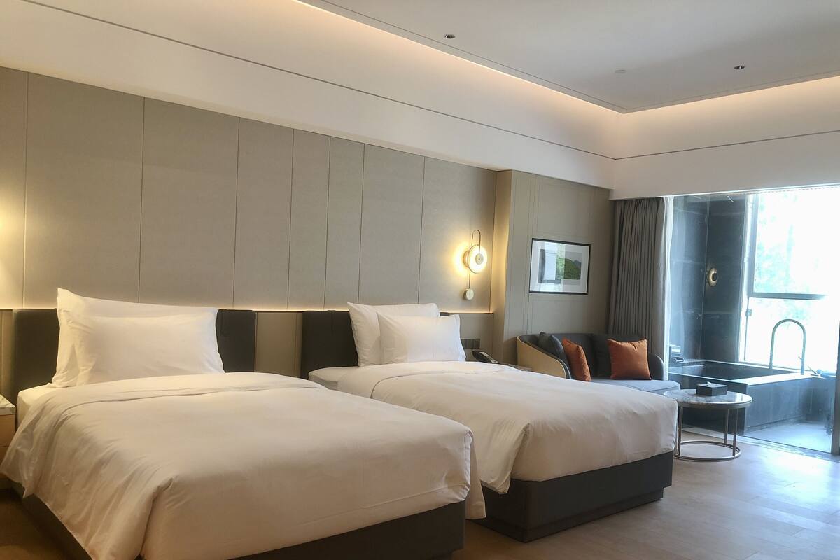 Una habitación de hotel moderna con dos camas, una zona de estar, un ventanal grande y cabina de ducha.