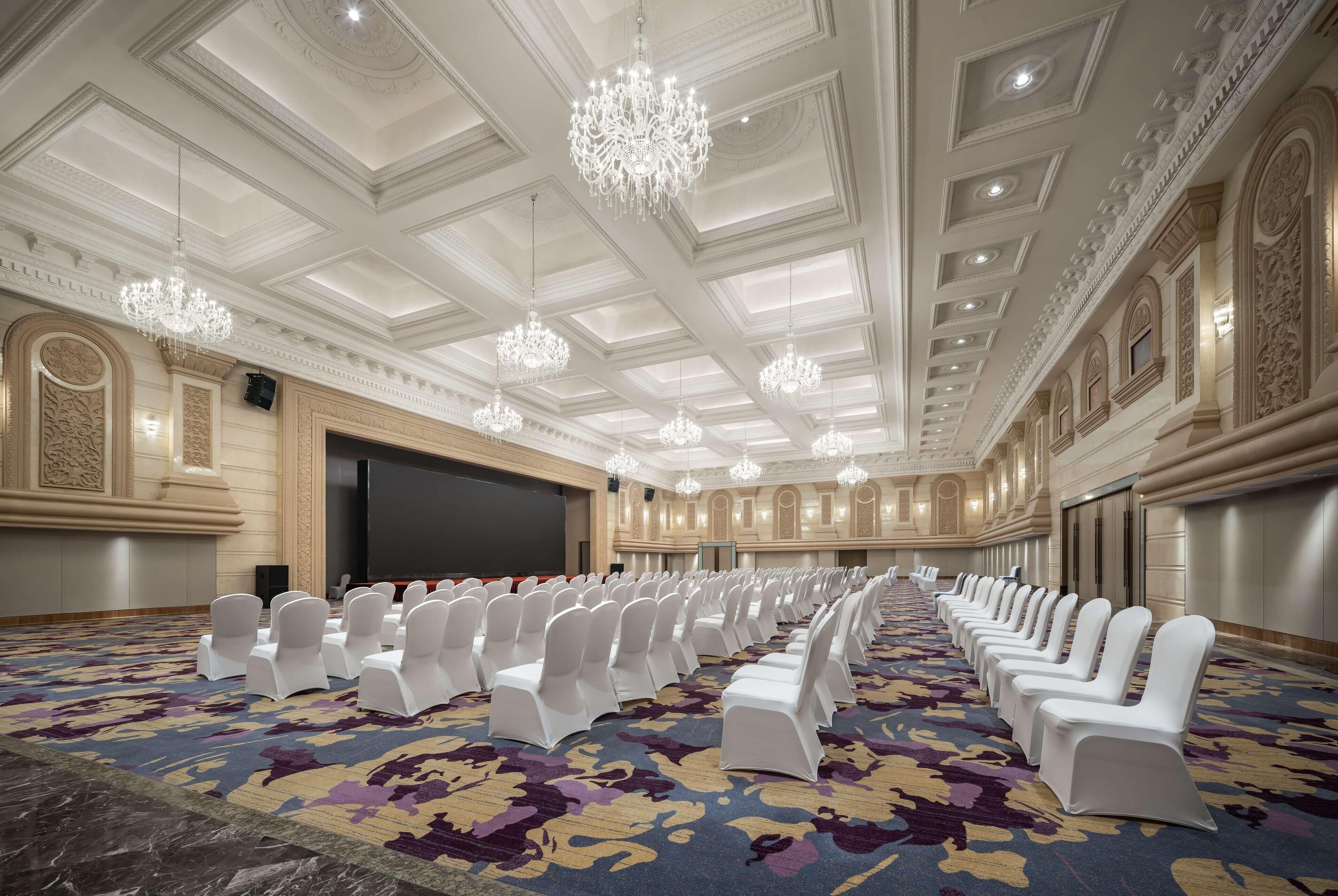 A conference room with rows of white chairs, a large screen, and ornate chandeliers.
