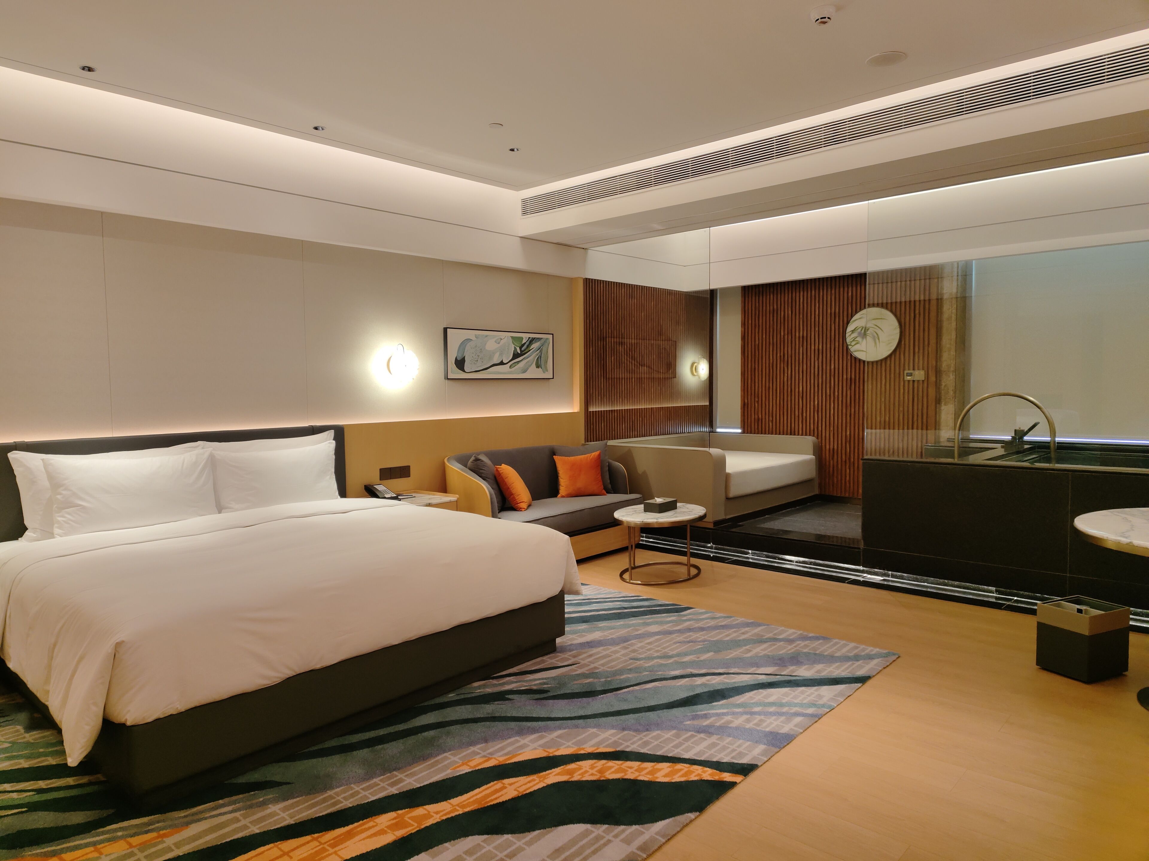 A modern hotel room with a large bed, a sofa, a small table, and a bathroom with a mirror and sink.