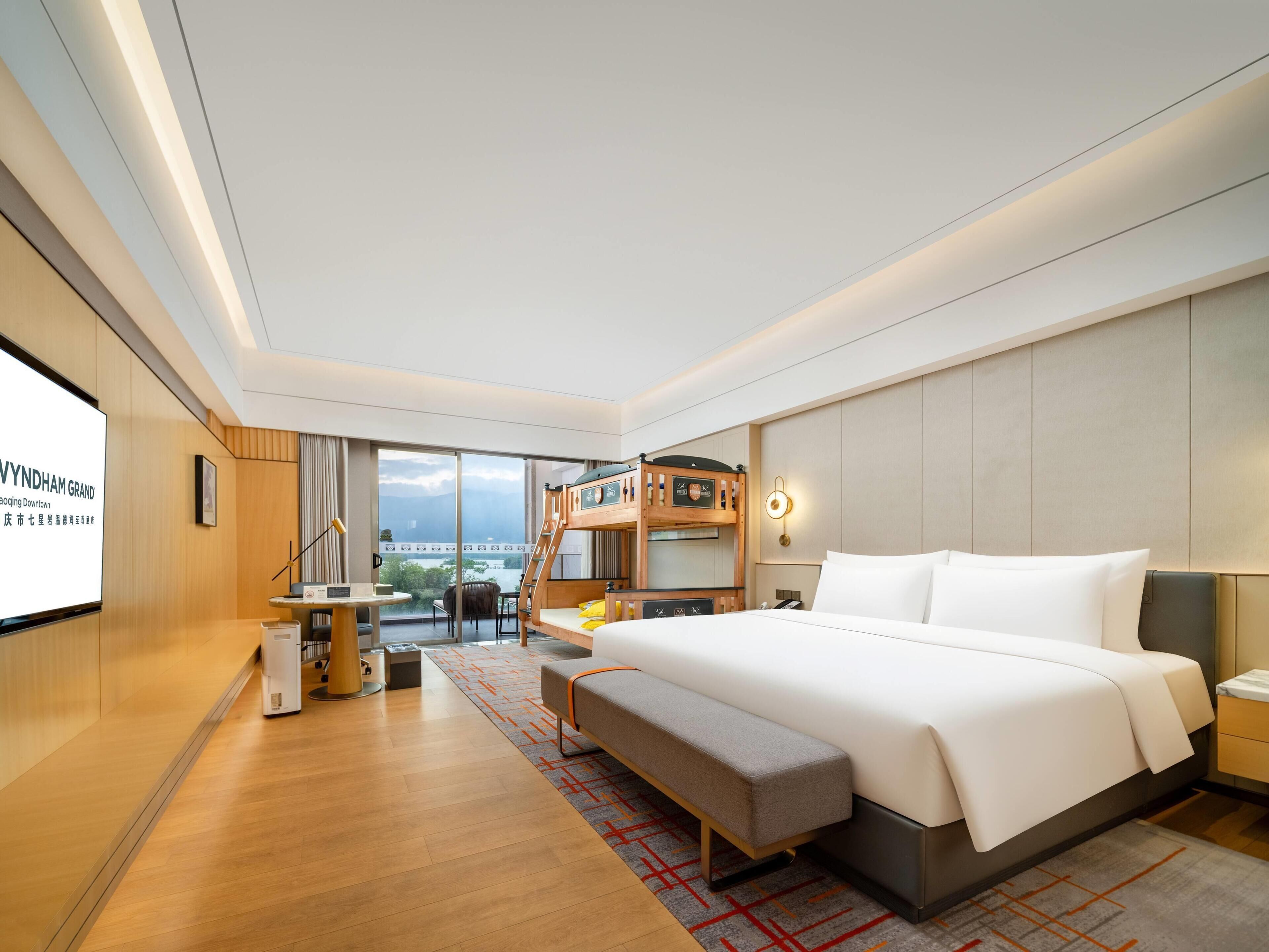 Una habitación de hotel moderna con una cama grande, un escritorio con silla, un pequeño rincón de descanso y un balcón con vistas a la ciudad.
